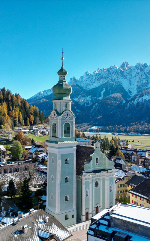 Light green church with tower, surrounded by houses, autumn-coloured trees and a snow-capped mountain
